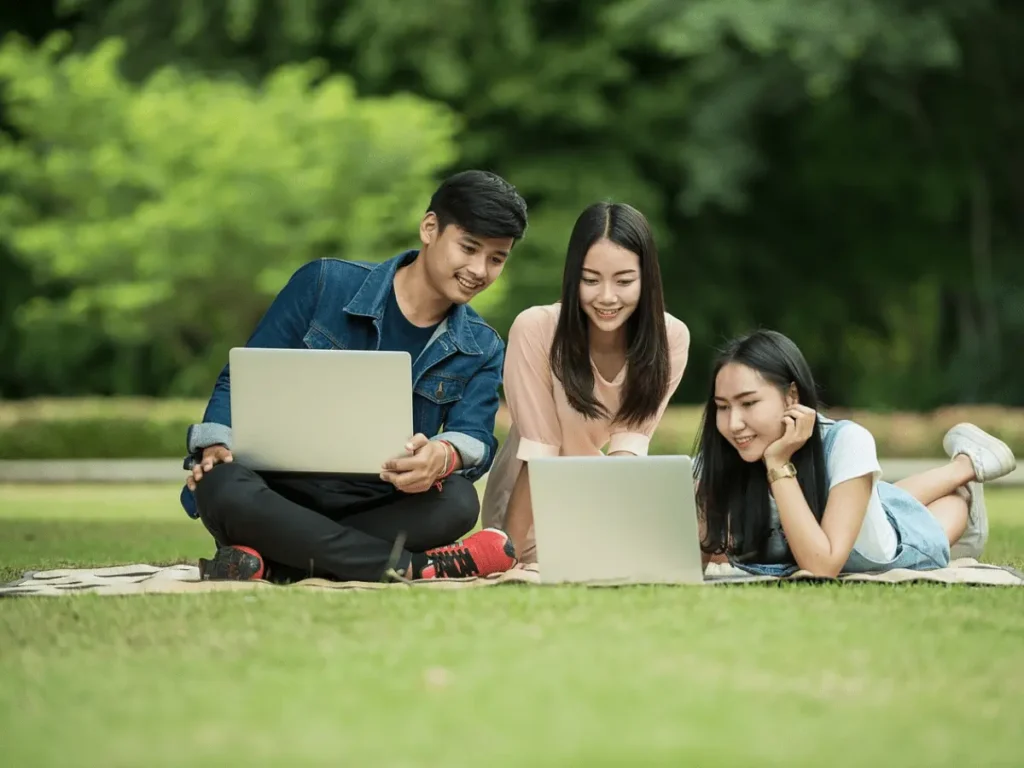 Three students are studying in the grass with two laptops